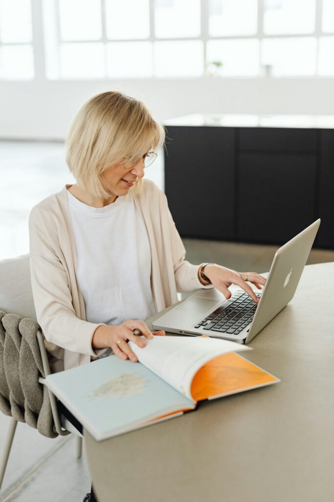 Caucasian woman with short blond hair working on a laptop and studying an open book indoors.